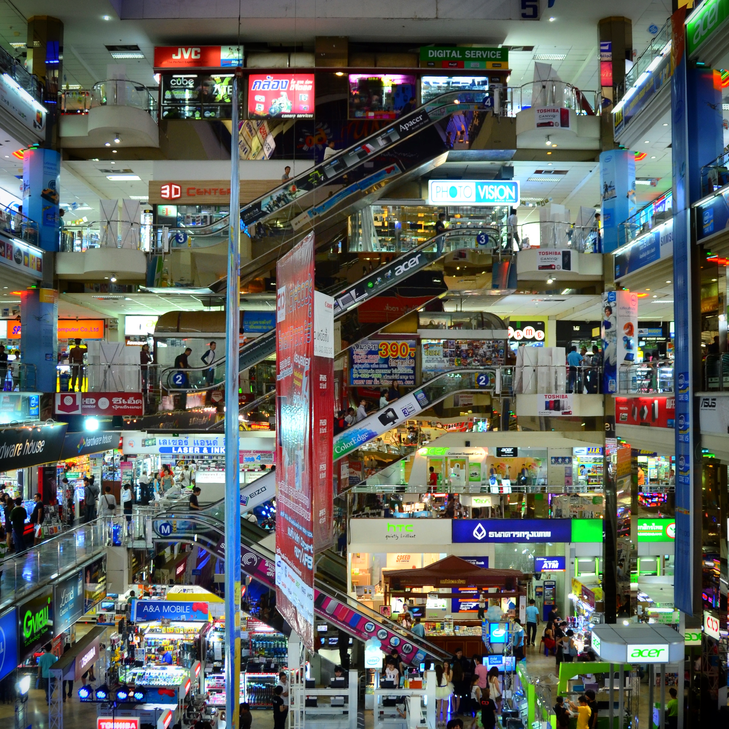Bustling multi-level shopping mall interior in Pratunam Market, Bangkok, with escalators, neon-lit shops, and crowds of shoppers